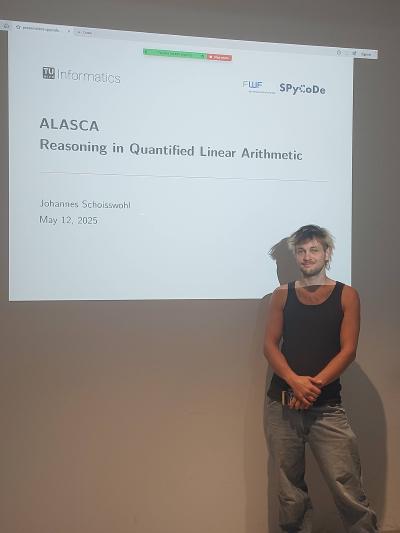 Johannes Schoisswohl is standing in front of a presentation screen that displays the title of the presentation and the logos of TU Wien Informatics, FWF and SPyCoDe.