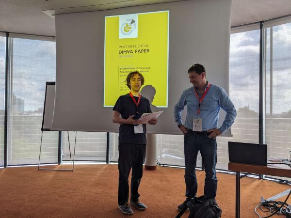 Two people stand in front of a projector screen displaying a yellow slide titled “Most Influential DIMVA Paper”