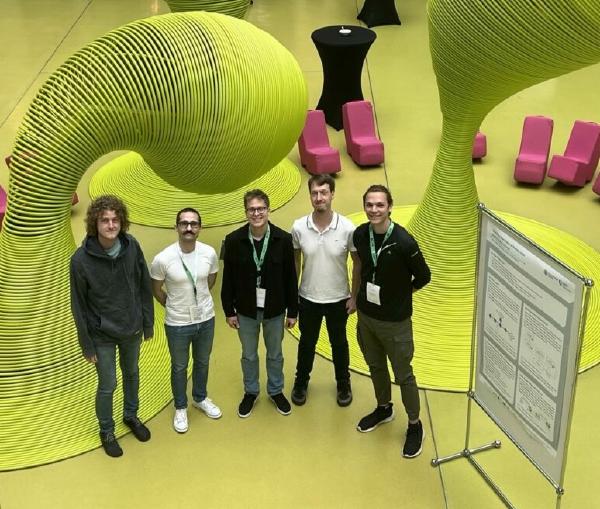 Five conference participants stand together smiling in a modern indoor space with a yellow floor and large abstract green sculptural elements in the background. There are poster presentations visible nearby.
